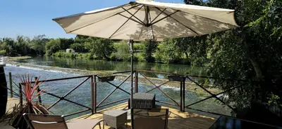 a patio with an umbrella and chairs next to a river at La terrasse du pêcheur in LʼIsle-sur-la-Sorgue